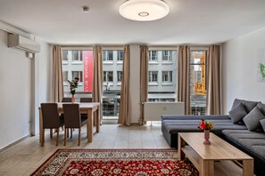 A modern living room with a sofa, a coffee table, a dining table with chairs, and a large window with curtains.