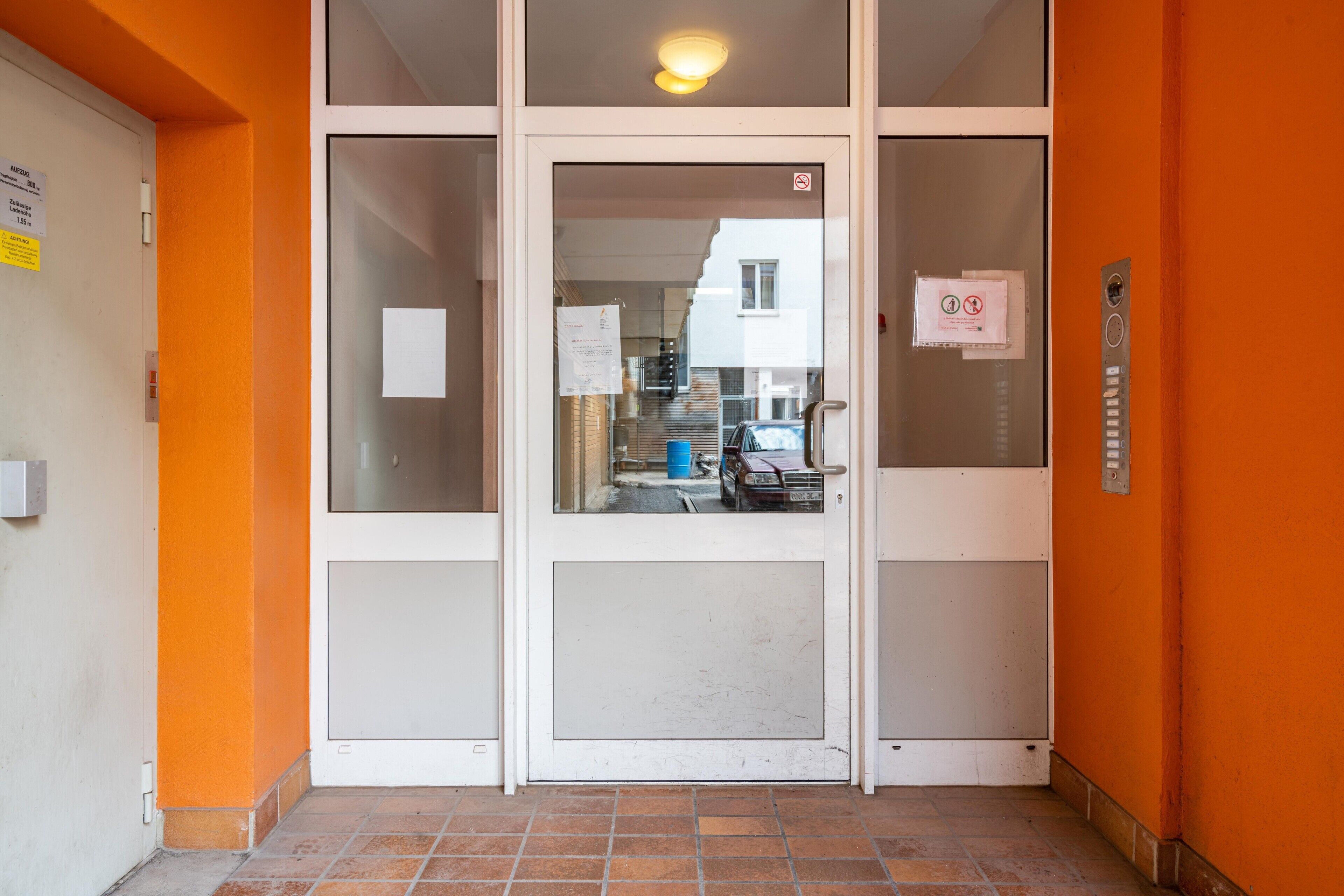 Entrada de un edificio con puerta de vidrio, un cartel de prohibido fumar y un detector de incendios en la pared.