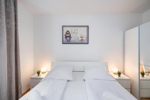 Un lit bien fait, avec du linge de lit blanc, deux tables de chevet avec des lampes, une œuvre d’art encadrée au mur et un grand miroir sur le côté droit.