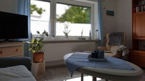A room with a television, a dining table with a blue tablecloth, a chair, a bookshelf, and a window with curtains.