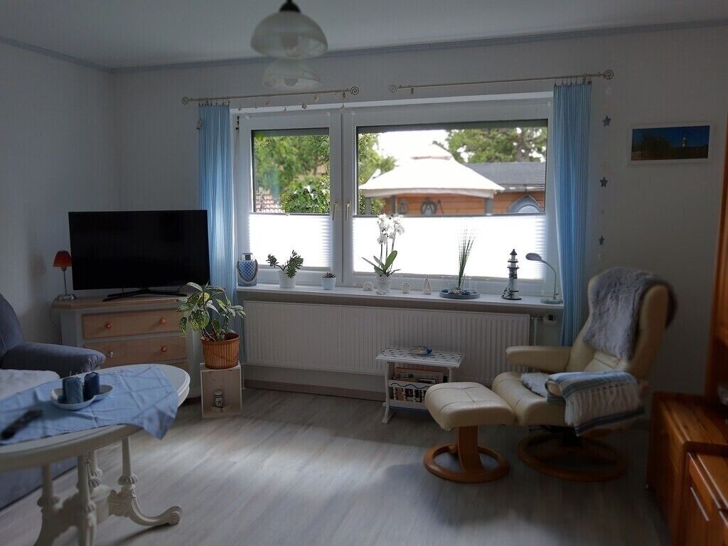 Ein Zimmer mit Fernseher, einem Sessel, einem kleinen Tisch, einer Bank und einem Fenster mit Vorhängen.