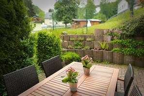 An outdoor wooden table with chairs, potted plants, and a stone retaining wall with greenery. - Apartment 'Fewo Malerstübchen' with Private Terrace and Wi-Fi (Willingen (Upland))