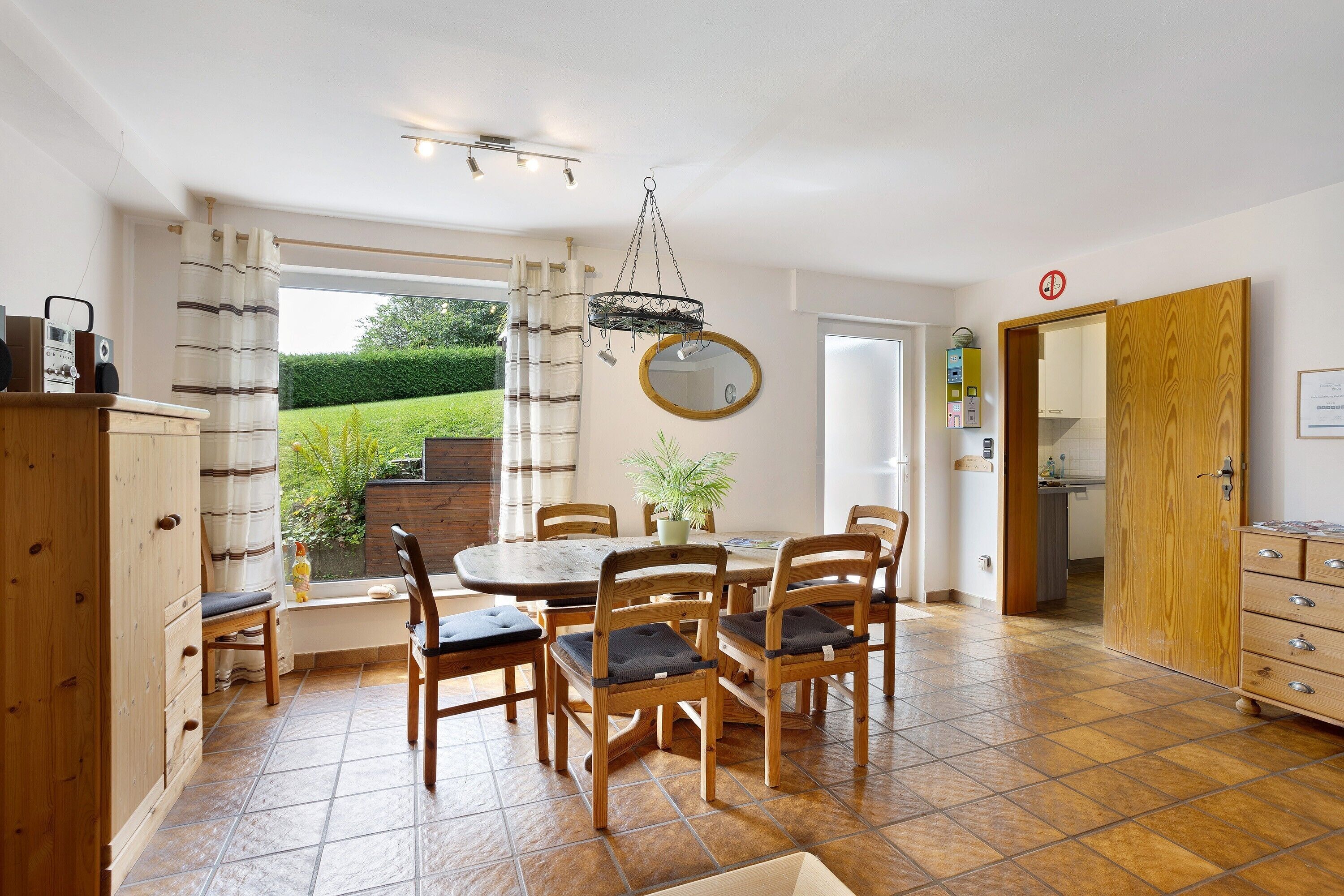 Un coin repas avec une table ronde, six chaises, un luminaire suspendu, une plante en pot et une vue sur un jardin par la fenêtre.