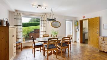 Un coin repas avec une table ronde, six chaises, un luminaire suspendu, une plante en pot et une vue sur un jardin par la fenêtre.