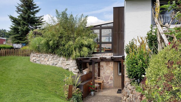 Een modern huis met een stenen gevel, een houten terras en een klein terras met een tafel en stoelen.
