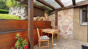 Een terras buiten met een houten tafel en stoelen, een stenen muur en een houten bank met rode bloemen.