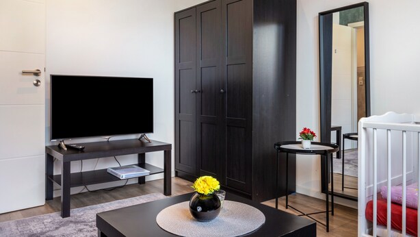 Un salon moderne avec un téléviseur à écran plat, une armoire en bois foncé, une petite table ronde ornée d’un vase de fleurs et un berceau à ossature blanche.