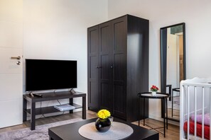 Een moderne woonkamer met een flatscreen televisie, een donker houten kledingkast, een klein rond tafeltje met een bloevas en een wieg met een wit frame.