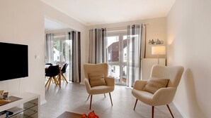 Ein modernes Wohnzimmer mit einem Flachbildfernseher, zwei beigen Sesseln, einem Glastisch und einem Essbereich mit Tisch und Stühlen.