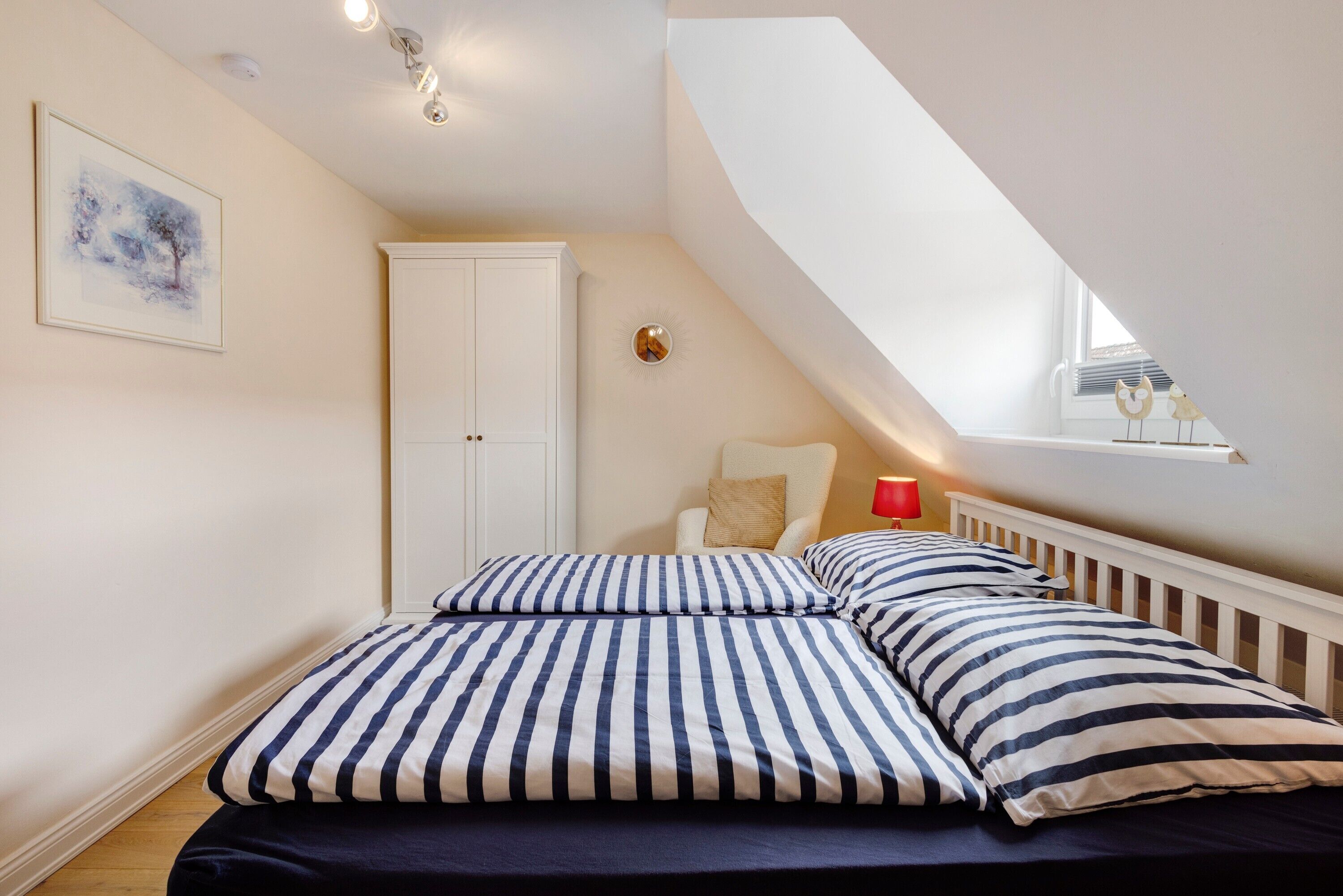 Ein Schlafzimmer mit schrägverdachter Decke, einem Bett mit gestreifter Bettwäsche, einem Sessel, einem kleinen Tisch mit einer roten Lampe und einem Fenster mit Dekoration.