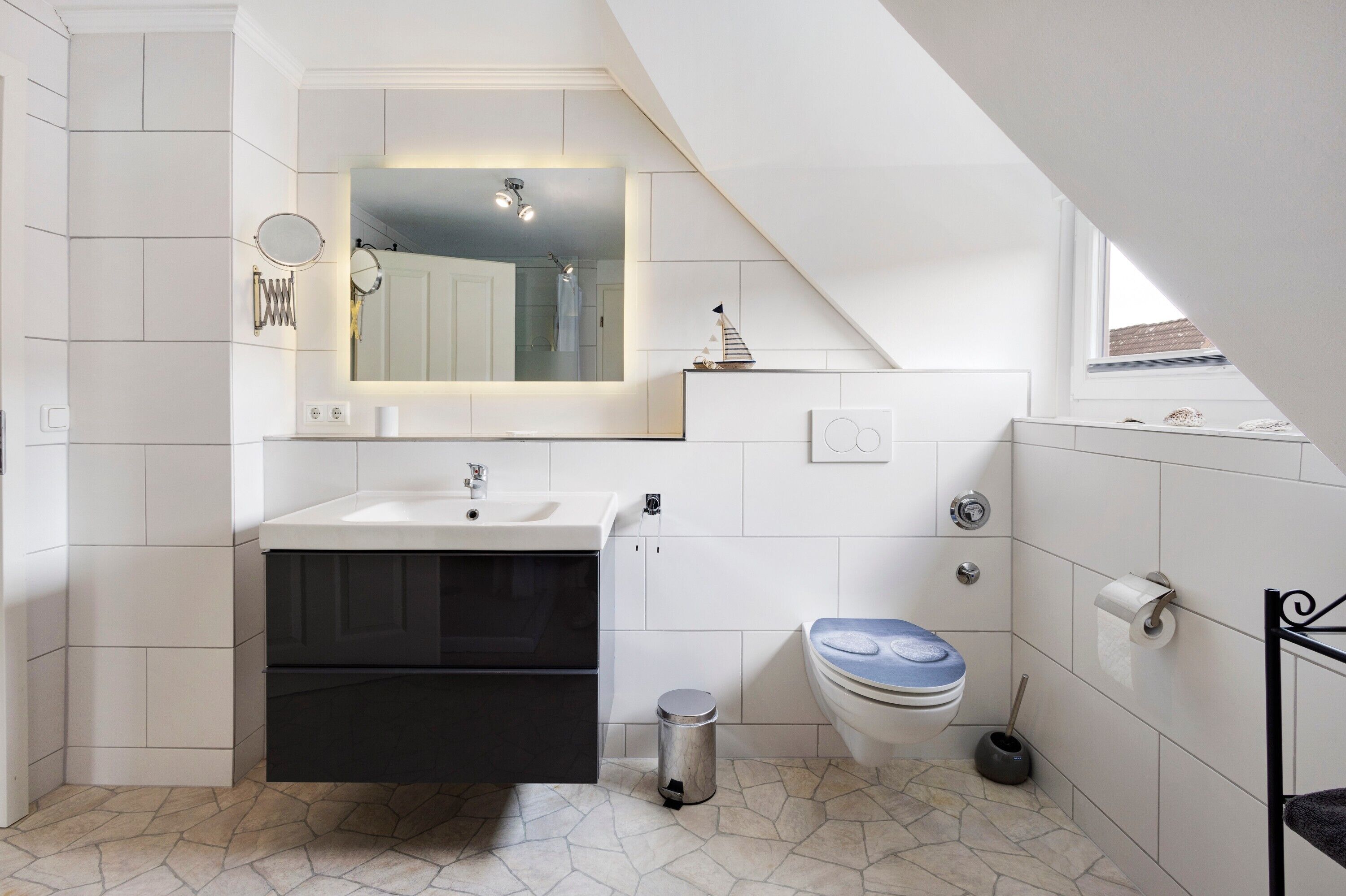 Un baño moderno con piso de baldosas blancas, un lavamanos con espejo arriba, un inodoro con tapa azul y un basurero pequeño.