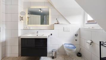 Un baño moderno con piso de baldosas blancas, un lavamanos con espejo arriba, un inodoro con tapa azul y un basurero pequeño.