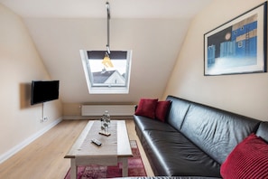 En moderne stue med en sort lædersofa, et træbord, et fjernsyn, et vindue med udsigt til en bygning og et indrammet billede på væggen.