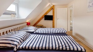 Ein Schlafzimmer mit schrägverdeckter Decke, einem an der Wand montierten Fernseher, einem Bett mit gestreifter Bettwäsche, einem kleinen runden Tisch und einer roten Lampe.