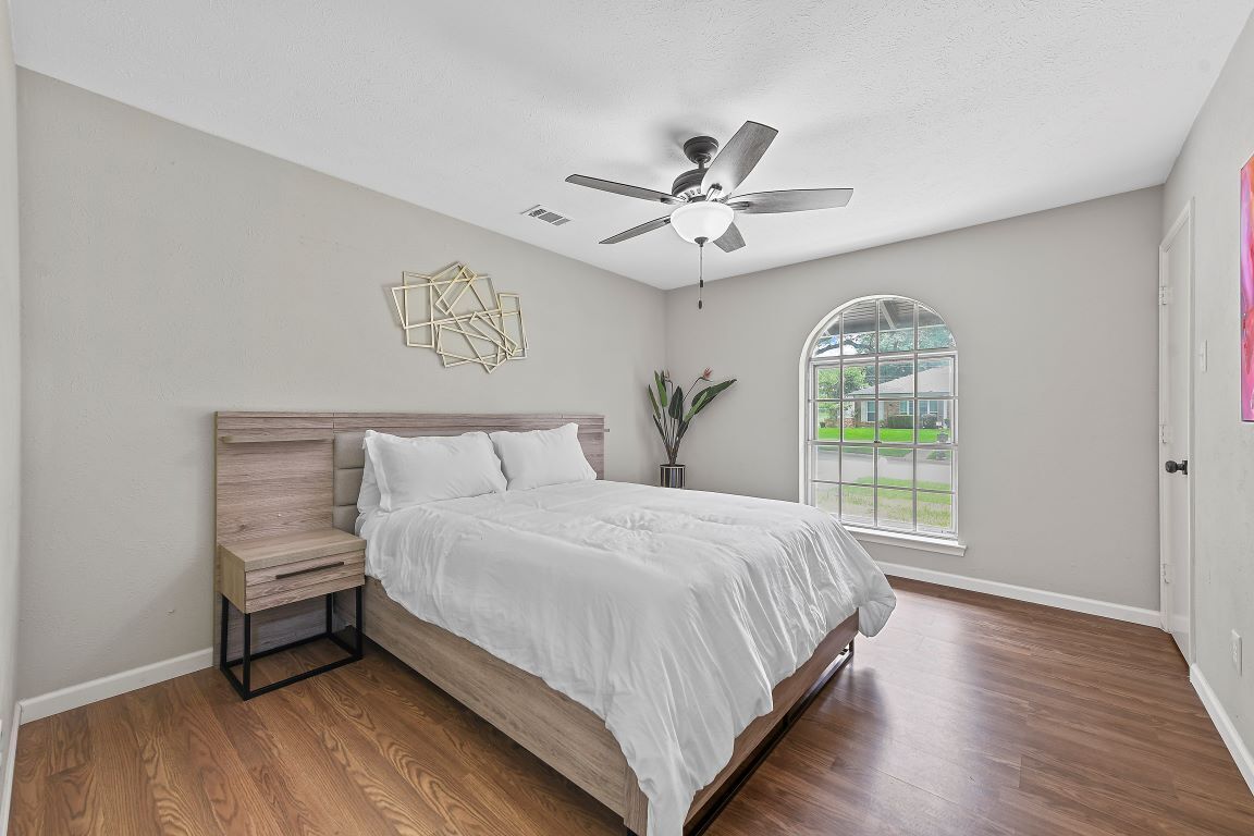 A bedroom with a bed, a nightstand, a ceiling fan, a window with a view, and a decorative wall piece.