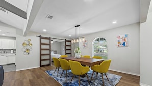 A dining area with a wooden table and yellow chairs, a blue patterned rug, and a kitchen in the background.