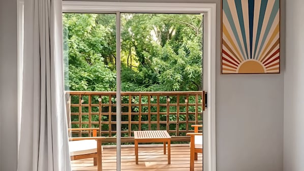 A balcony with a wooden table and bench, a view of lush greenery, and a sunburst artwork on the wall.