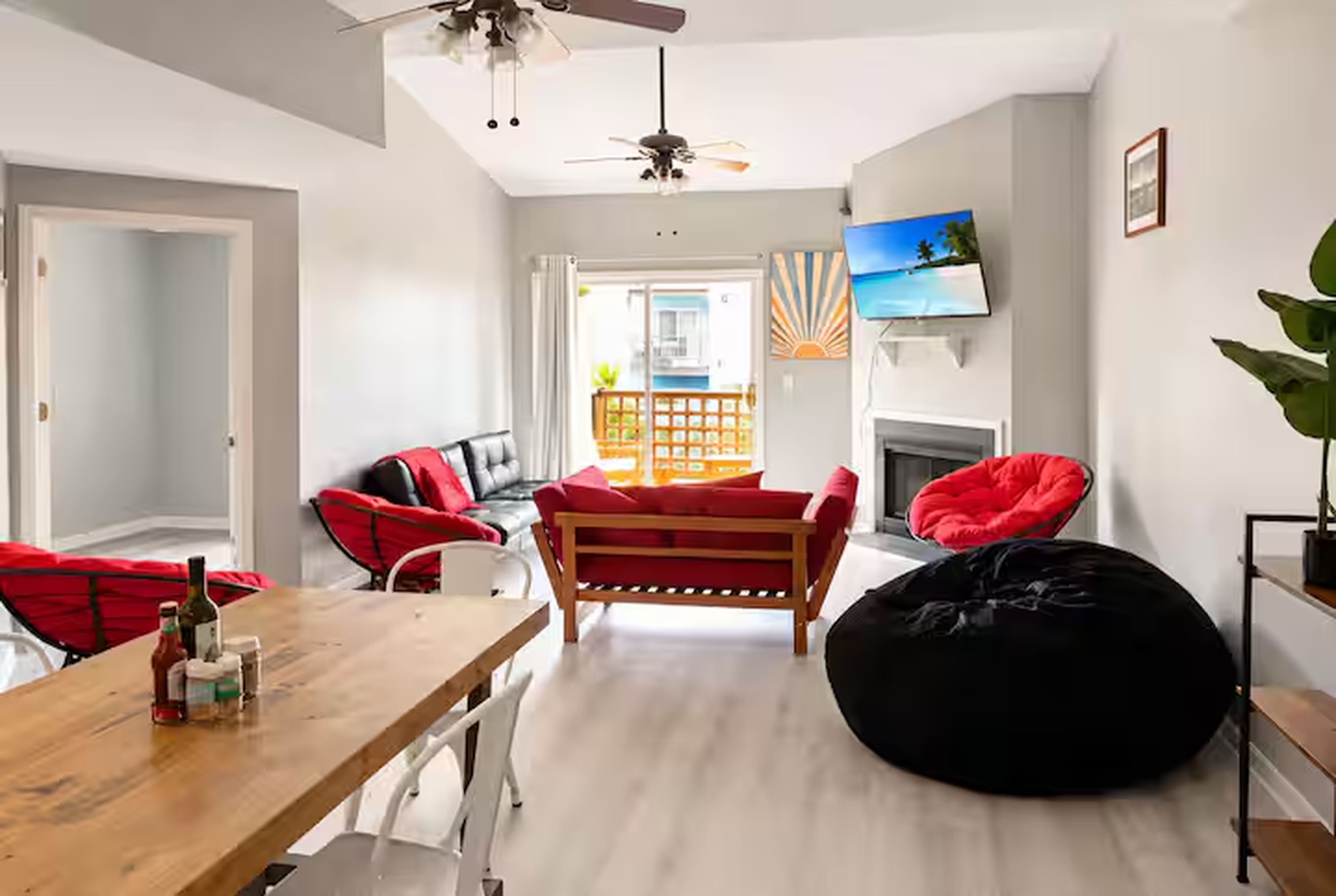 A modern living room with a wooden table, red chairs, a black bean bag, a ceiling fan, and a TV on the wall.