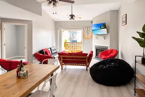 A modern living room with a wooden table, red chairs, a black bean bag, a ceiling fan, and a TV on the wall.