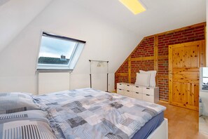 Une chambre avec un plafond en pente, une fenêtre, un lit recouvert d’une couvre-lit à motifs, une armoire en bois et un radiateur.
