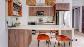 A modern kitchen with wooden cabinets, a central island with bar stools, a hanging light fixture, and a stainless steel refrigerator. - TAO Penthouse, Private Rooftop SpaTub, Pool Onsite (Tulum)