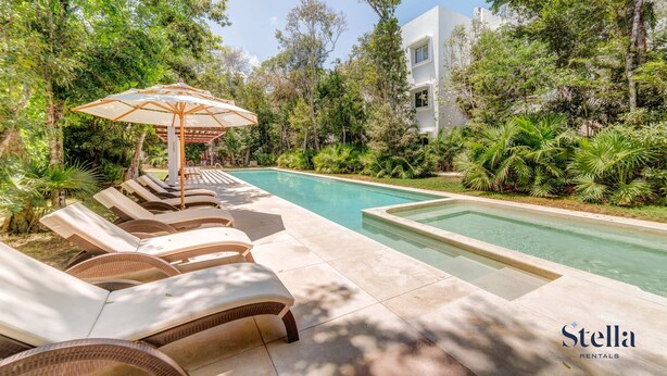 A swimming pool area with lounge chairs and an umbrella, surrounded by lush greenery and a modern building. - TAO Penthouse, Private Rooftop SpaTub, Pool Onsite (Tulum)
