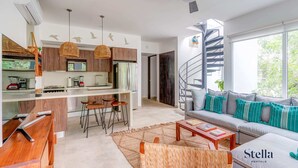 A modern living room with a sofa, coffee table, and bar stools. There is a kitchen area with wooden cabinets and a dining table. A spiral staircase leads to an upper level. - TAO Penthouse, Private Rooftop SpaTub, Pool Onsite (Tulum)