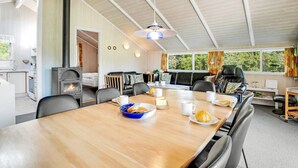 A modern, well-lit room with a wooden dining table, comfortable seating, a fireplace, and large windows offering a view of greenery outside. - "Guja" - 250m from the sea by Interhome (Faxe Ladeplads)