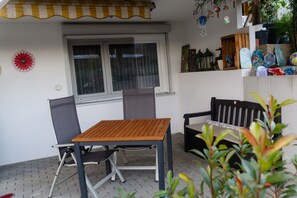 Une terrasse extérieure avec une table et des chaises en bois, une fenêtre avec des rideaux et une étagère ornée d’objets décoratifs.