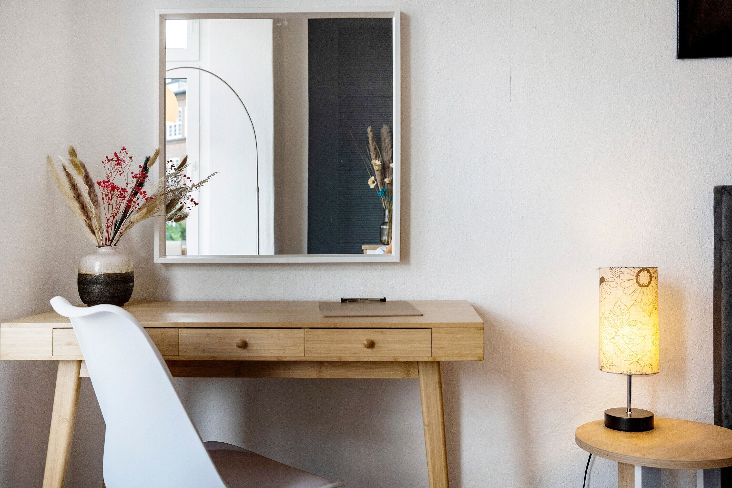 A wooden desk with a mirror, a vase with dried flowers, and a table lamp.