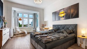 Ein Schlafzimmer mit einem großen Bett, einem Sessel, einem Schreibtisch, einem Fenster mit Vorhängen und einer Chandelier.