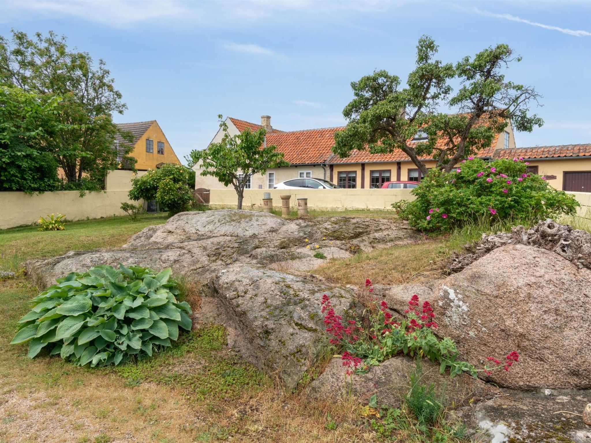 Ett bostadsområde med stora stenar, en trädgård med gröna växter och rosa blommor samt hus med rödfärgade tegeltak.