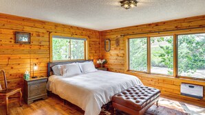 A bedroom with a wooden log wall, a large bed with white linens, a wooden nightstand, a leather bench, and a cowhide rug. - Dubois Riverfront Lodge w/ On-Site Fishing & Views (Dubois)