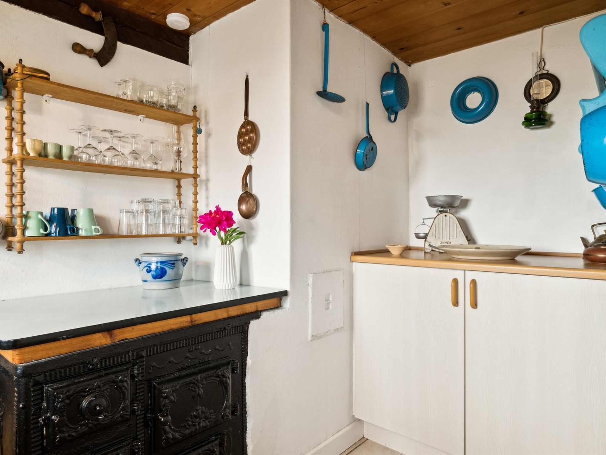 A kitchen with wooden shelves holding glassware and mugs, a hanging pot rack, and a vintage scale on the counter.