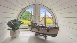 Un modelo de barco sobre una estantería con una planta en maceta al lado, visto a través de una ventana con un paisaje rural.