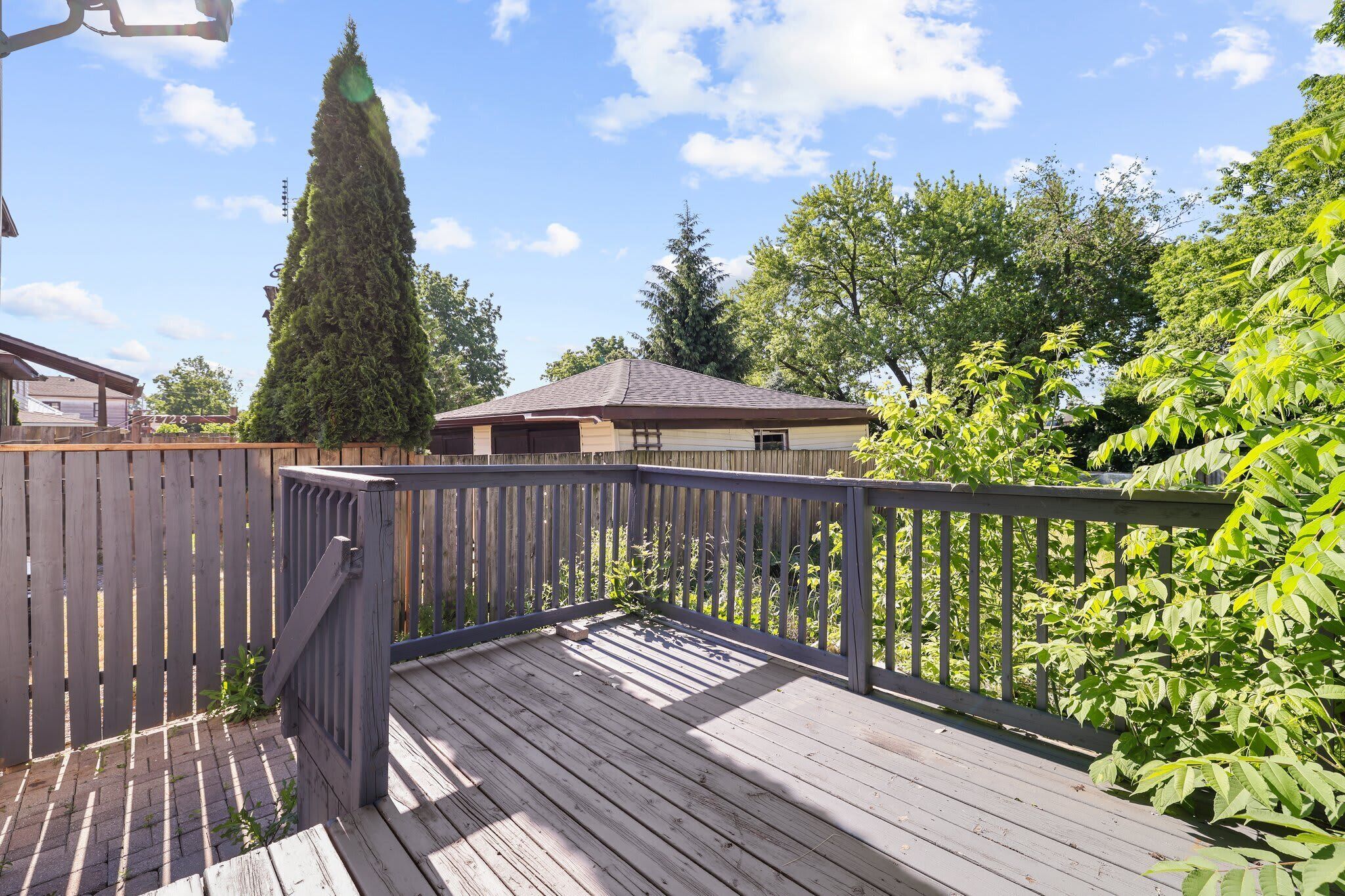 A wooden deck with a railing, surrounded by greenery and a house in the background.