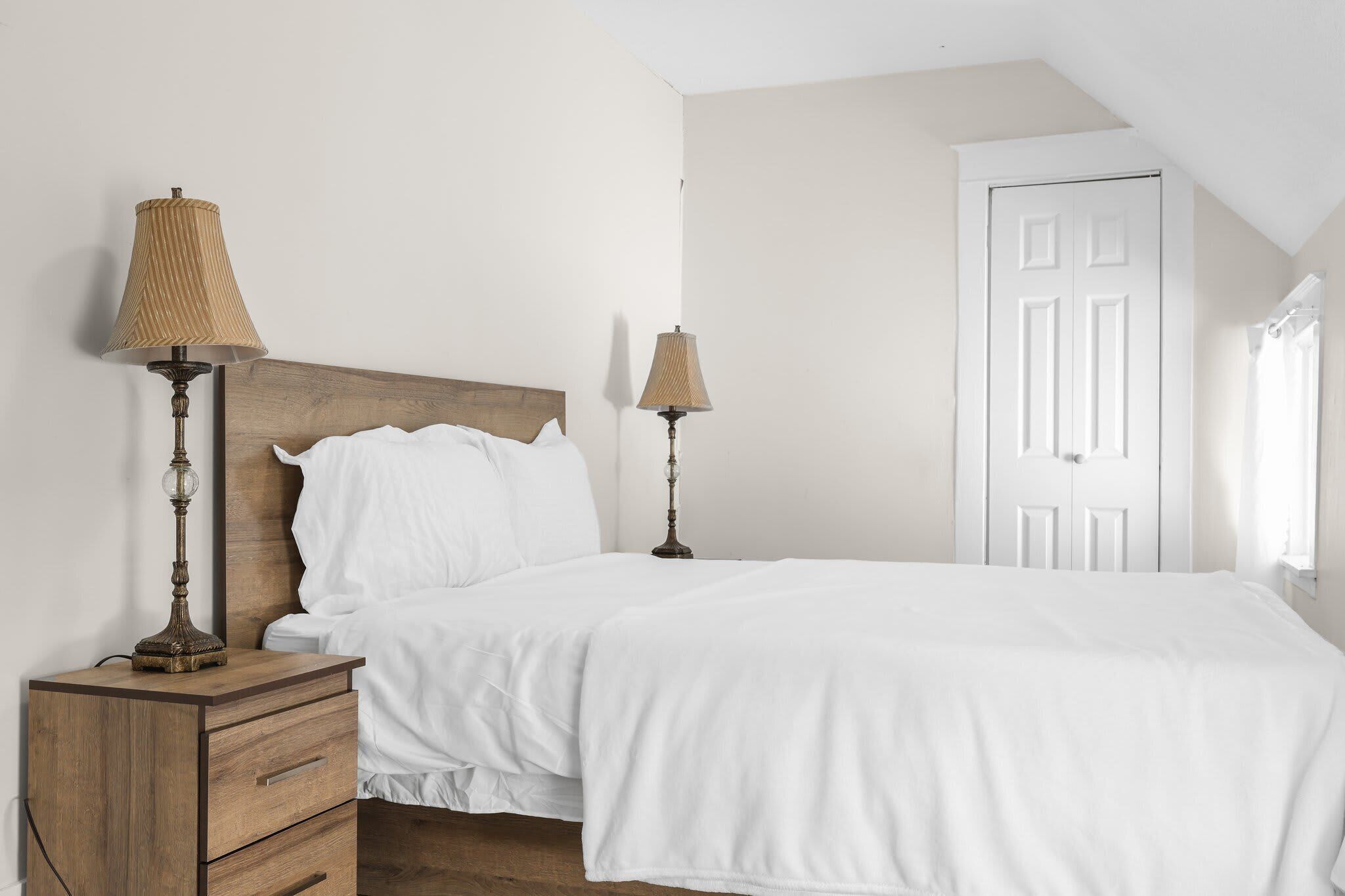 Un dormitorio con cabecera de madera, dos lámparas de noche, una colcha blanca, una mesita de noche con cajones y una puerta blanca.