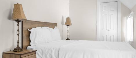 Un dormitorio con cabecera de madera, dos lámparas de noche, una colcha blanca, una mesita de noche con cajones y una puerta blanca.
