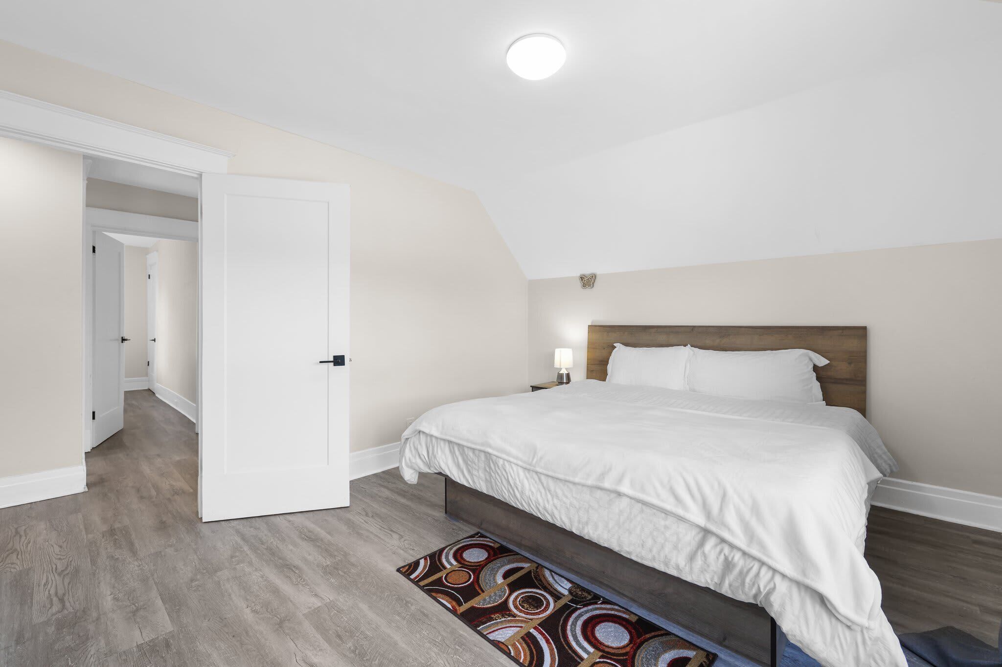 A bedroom with a large bed, a wooden headboard, a nightstand with a lamp, and a patterned rug.
