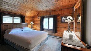 Un dormitori de cabana de fusta amb un llit, dues finestres, una tauleta de nit i una làmpada.