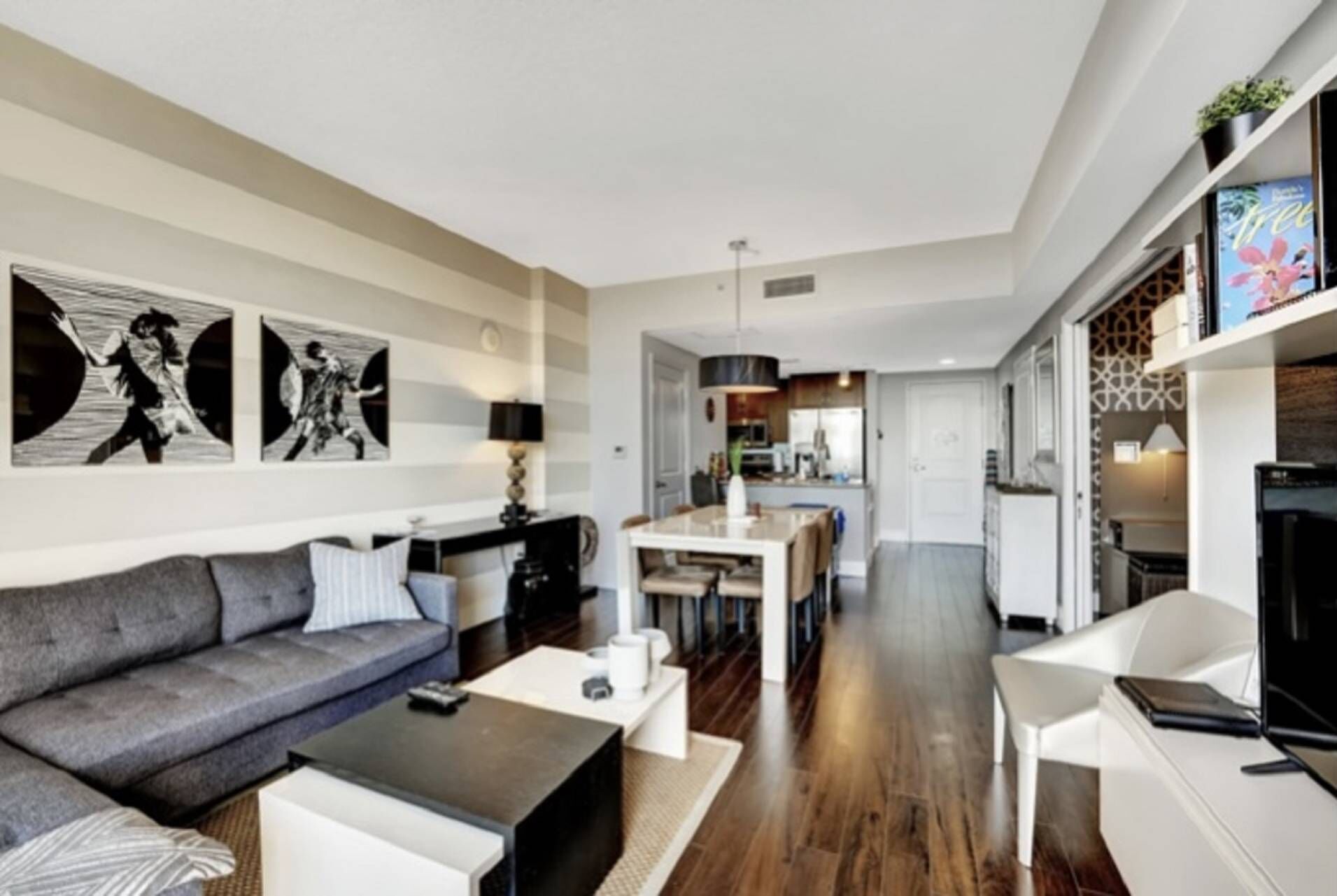 Un salón moderno con un sofá gris, una mesita de centro y un comedor con una mesa y sillas blancas. Se ve una cocina al fondo.