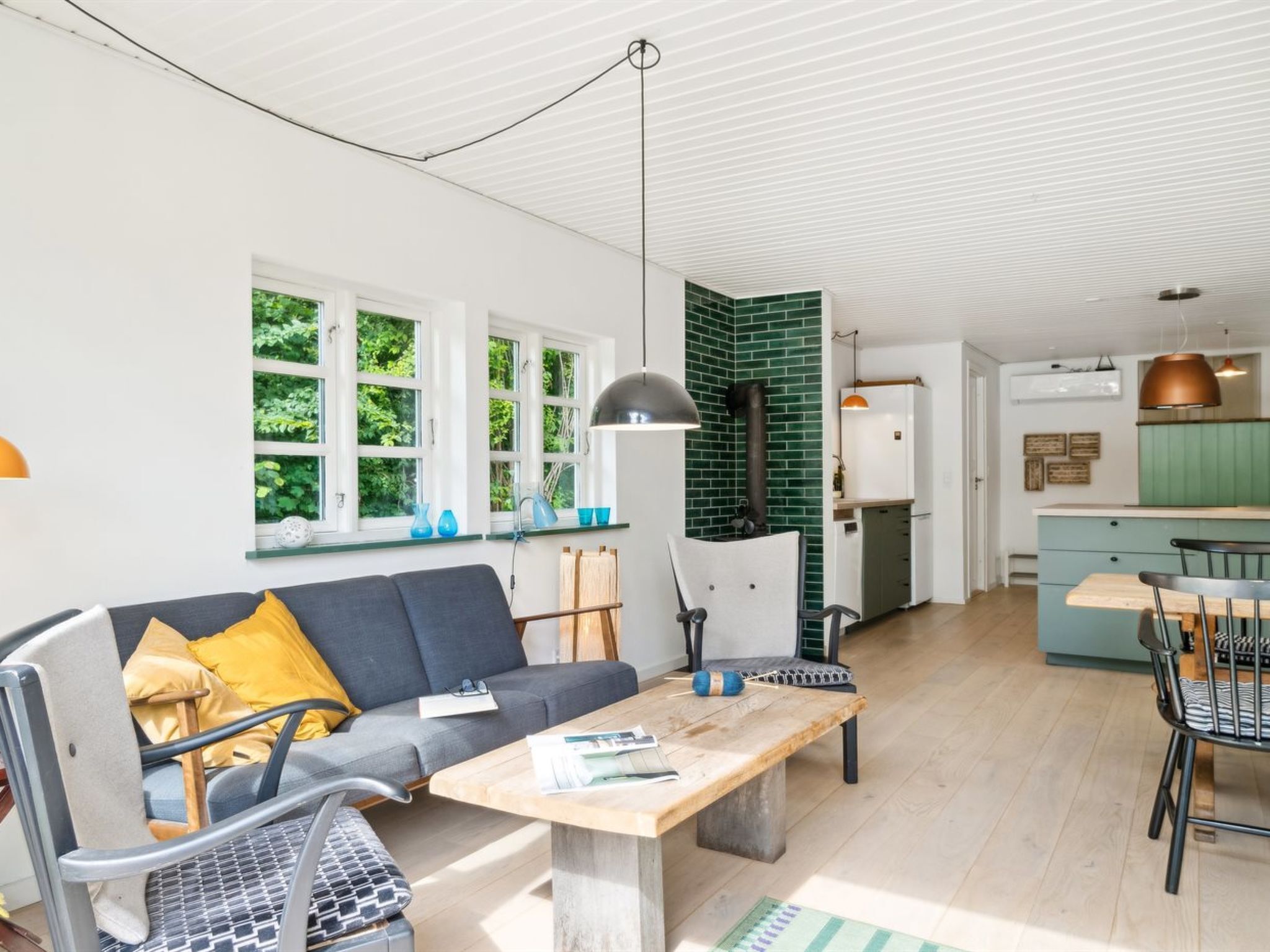 Ein modernes Wohnzimmer mit Sofa, Couchtisch und Sesseln, eine Küche mit Herd und große Fenster mit Blick auf Grünflächen.