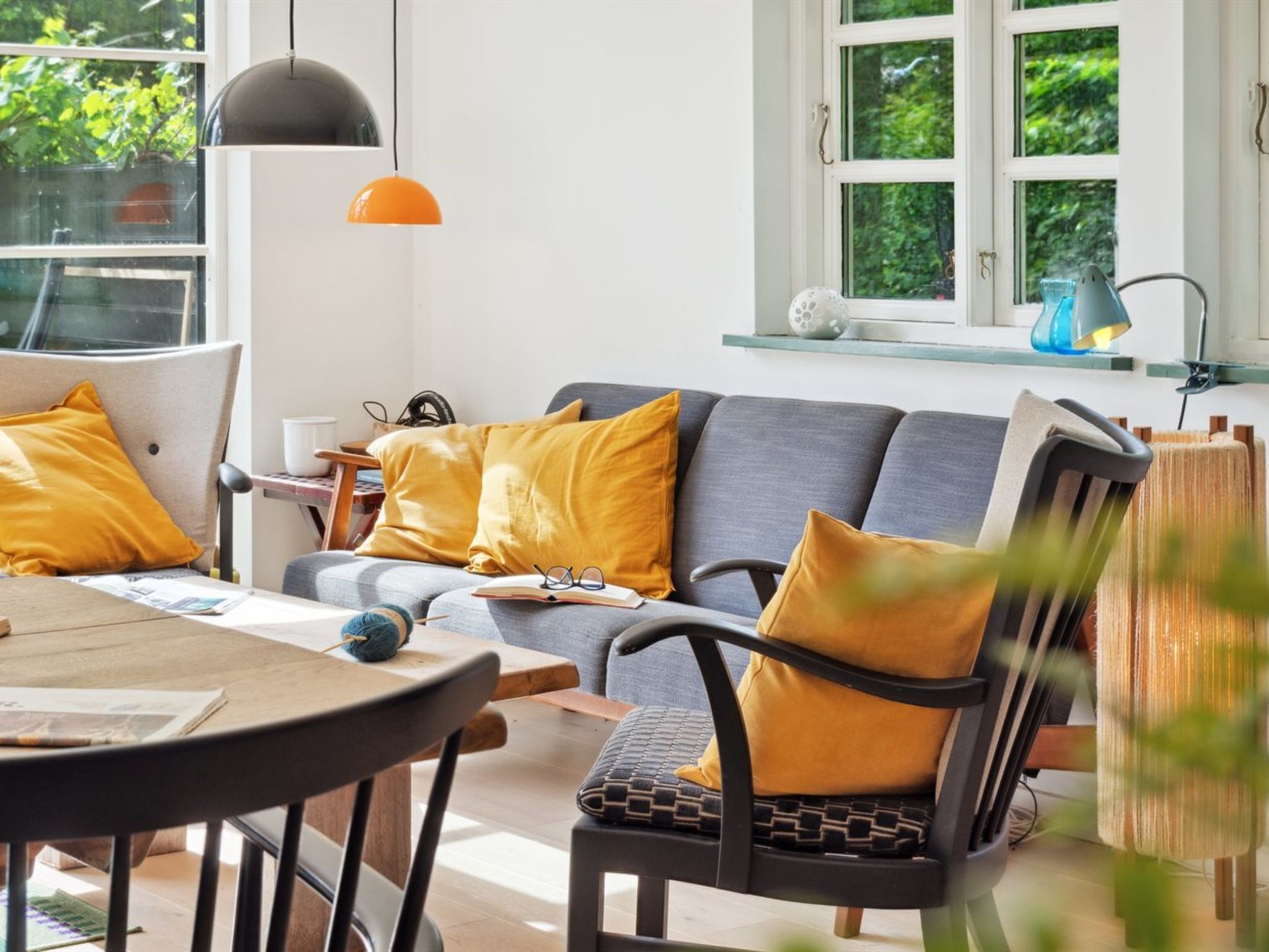 Ein modernes Wohnzimmer mit einer grauen Couch, gelben Kissen, einem runden Holztisch und einem Fenster mit Grünpflanzen draußen.