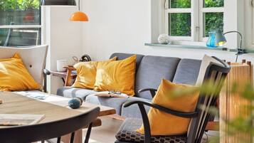 Un salon moderne avec un canapé gris, des coussins jaunes, une table ronde en bois et une fenêtre donnant sur un espace verdoyant.