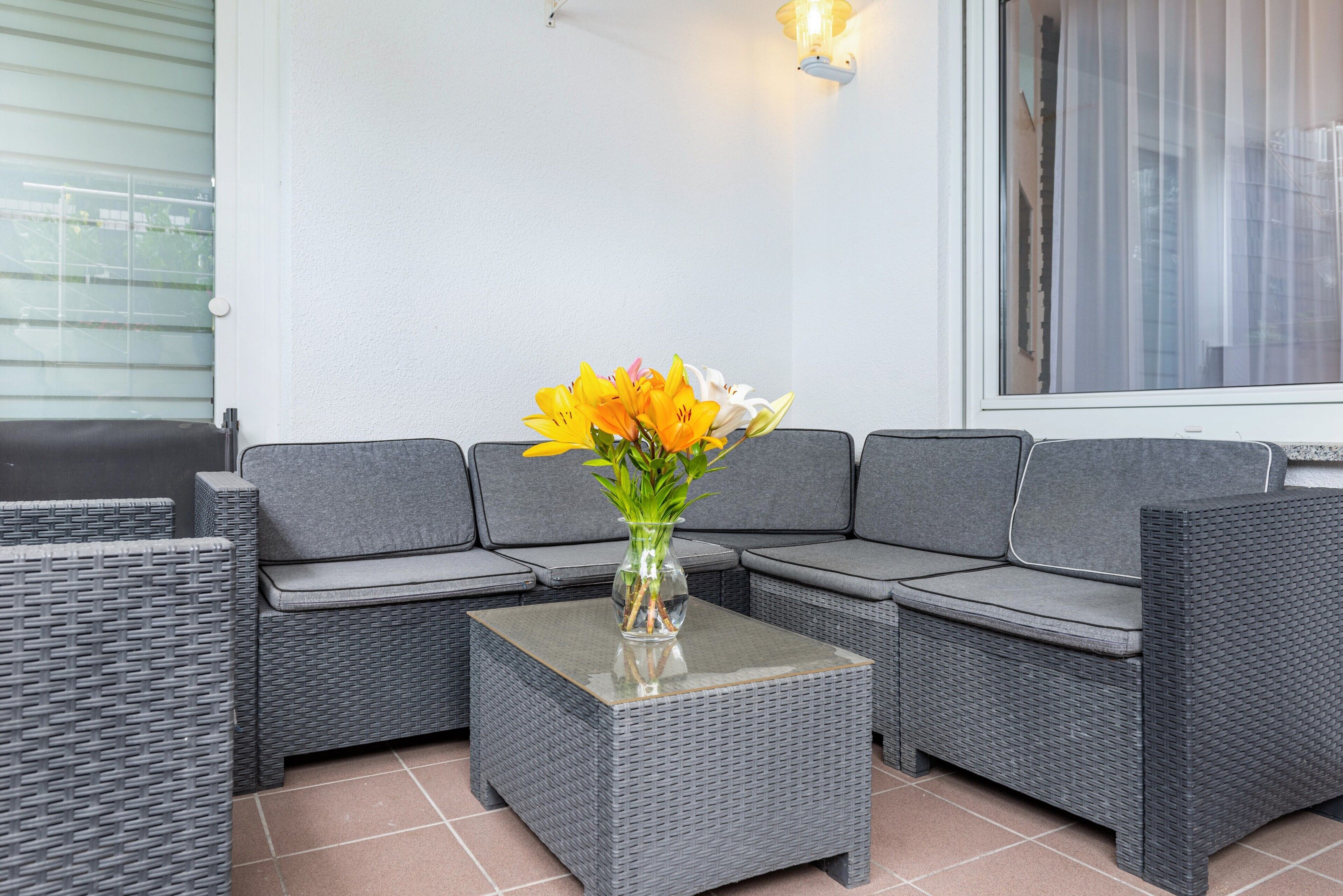 Eine graue Korbusitzgruppe mit einem Couchtisch mit Glasplatte und einer Vase mit Blumen.