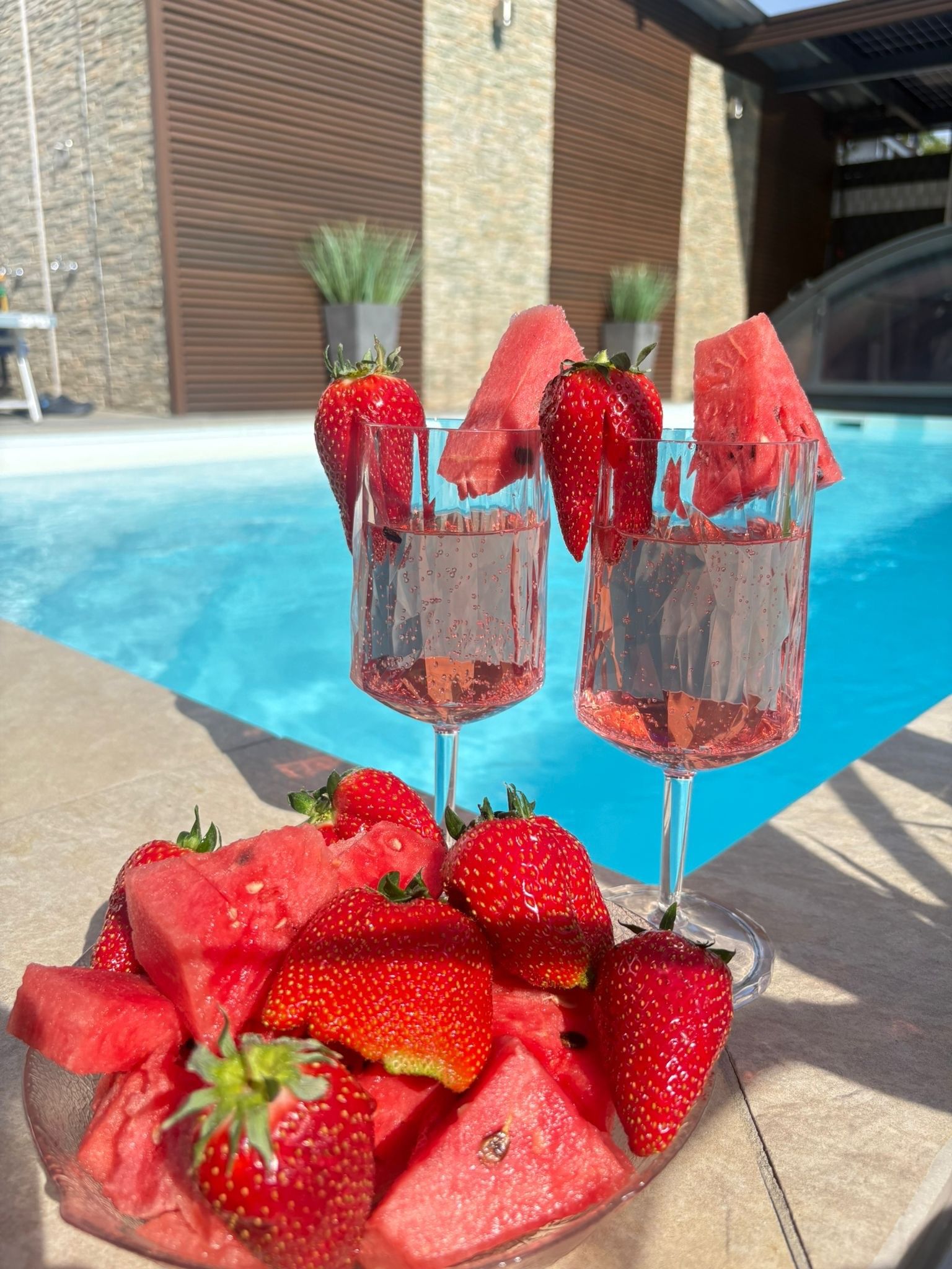 Une scène au bord de la piscine avec deux verres d’une boisson rose, une assiette de fraises et de pastèque, et un mur de pierre en arrière-plan.