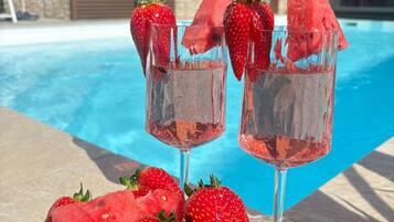 Une scène au bord de la piscine avec deux verres d’une boisson rose, une assiette de fraises et de pastèque, et un mur de pierre en arrière-plan.