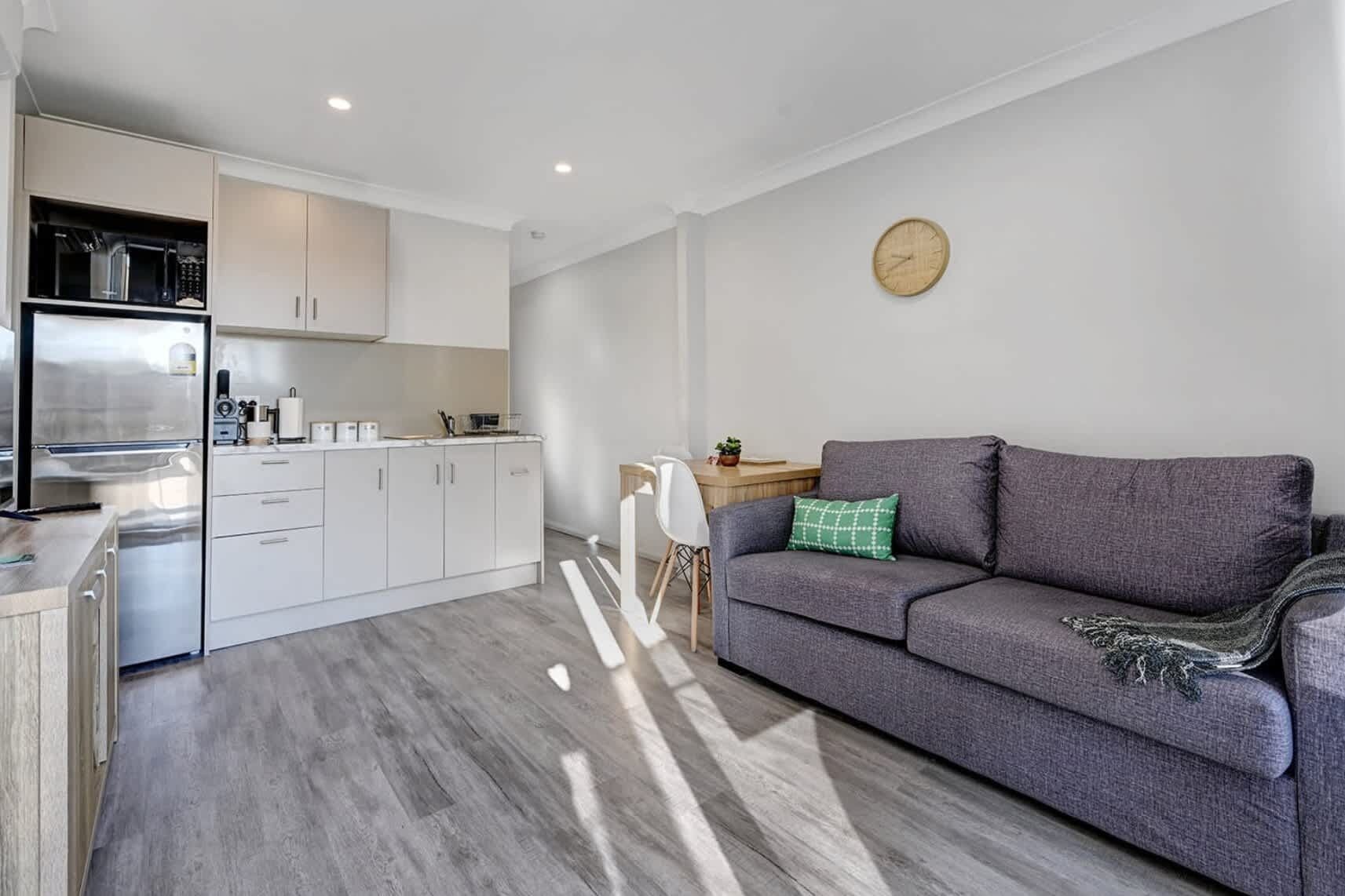 A modern kitchen with white cabinets and stainless steel appliances, a grey sofa with a green pillow, and a wooden dining table with chairs.