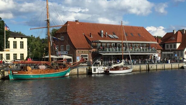 A harbor with boats docked, a building with a red roof, and a sign that reads 'EM. Scheel.'