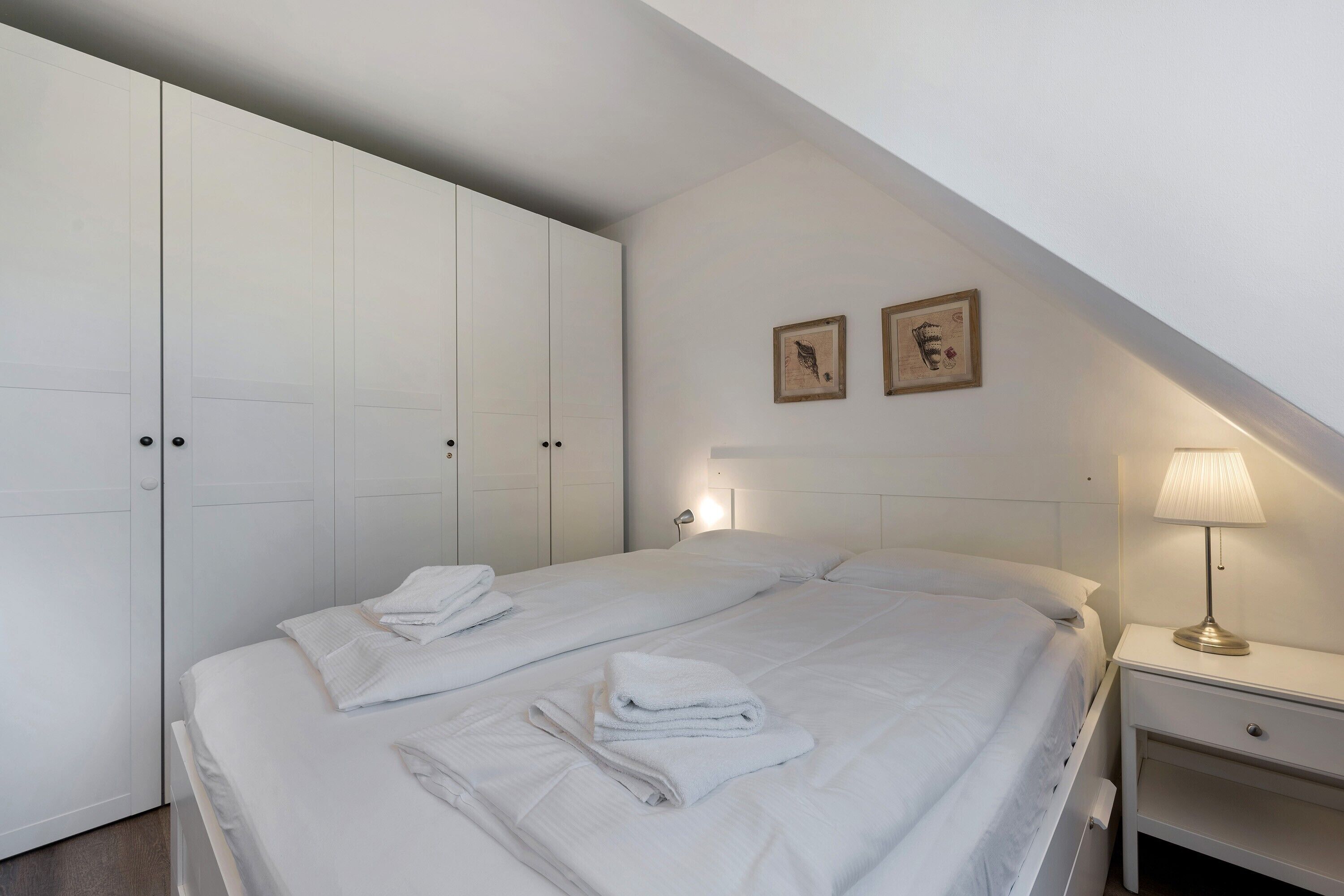 Ein Schlafzimmer mit einem Bett, zwei Nachttischen, einer Lampe und zwei gerahmten Bildern an der Wand.